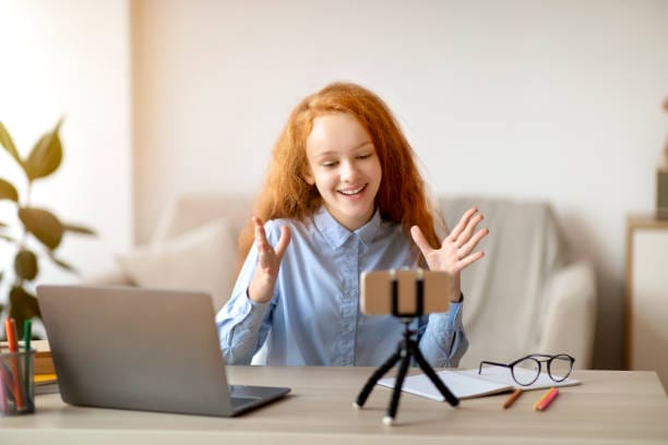 Blogging Concept. Smiling red-haired teen girl recording lifestyle blog talking to camera of mobile phone on tripod, sitting at desk at home. Happy young influencer filming vlog for her channel